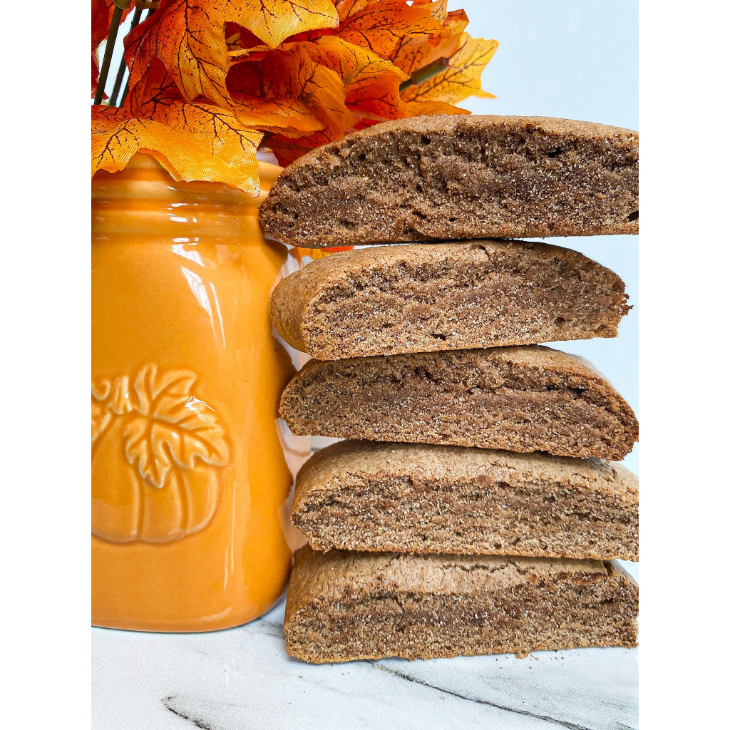 5 pieces of Pumpkin Spice Mandel Bread, shaped like biscotti, but chewy like a cookie & with the color of gingerbread, all stacked on a marble table with an orange painted mason jar with a pumpkin carving filled with leaves.